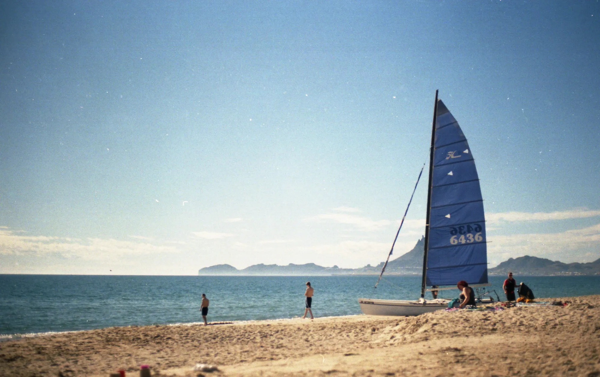 A small sailboat framed by pale water, beach, and distant hills.