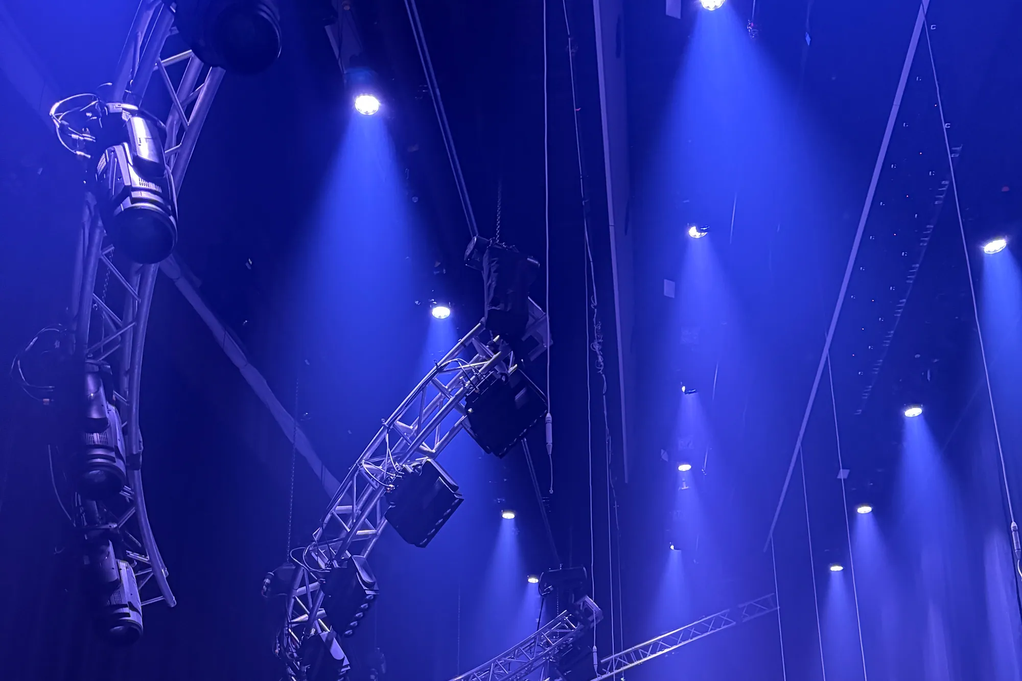 Haze making the light geometry visible — the audience sees atmosphere, the production team sees angles, timing, fixtures, power, and a lot of cable.